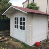 Una casetta in legno con porte di vetro e un tetto rosso situata in un giardino con un mucchio di legna accatastato vicino
