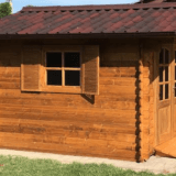 Una piccola casetta in legno con tetto in tegole e finestre dotate di persiane, circondata da verde